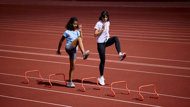 Teenage female runner practicing high knee hurdle drill beside adult female coach. Concept of agility and speed training, junior hurdle practice, summer sport coaching.