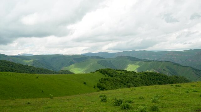 A panoramic view of a rolling foothill landscape: in the foreground are gentle grassy slopes dotted with sparse trees and patches of wildflowers; in the middle zone are rounded hills with dense green 
