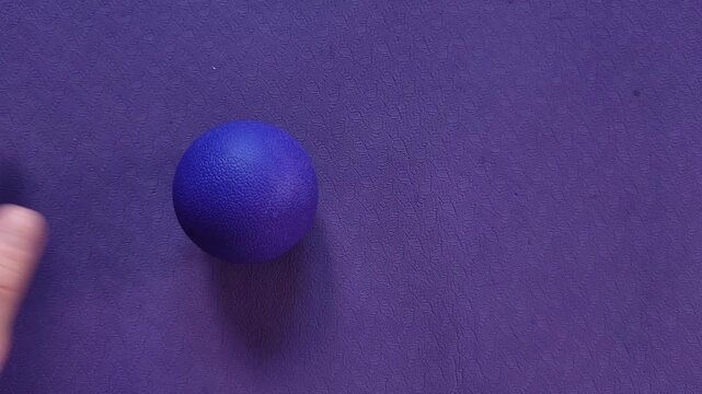 A woman places a small purple massage ball on a purple exercise mat, self massage practice, wellness routine, foot care.