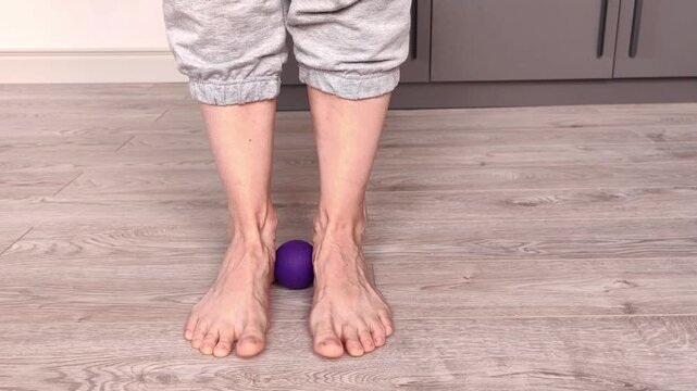 An adult woman squeezes a massage ball between her ankles and rises onto her tiptoes, practicing foot massage and balance exercise at home.