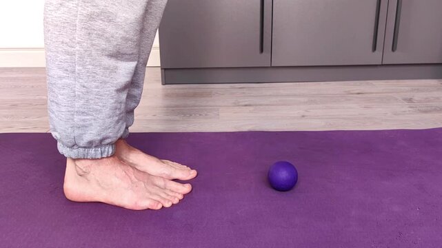 An adult woman rises onto her tiptoes next to a small purple massage ball on an exercise mat, foot mobility, self massage, wellness routine.