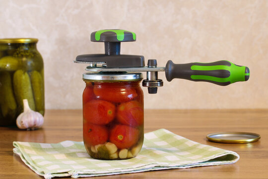 Manual canning sealer closing glass jar with homemade preserved tomatoes, garlic and vegetables on kitchen table.