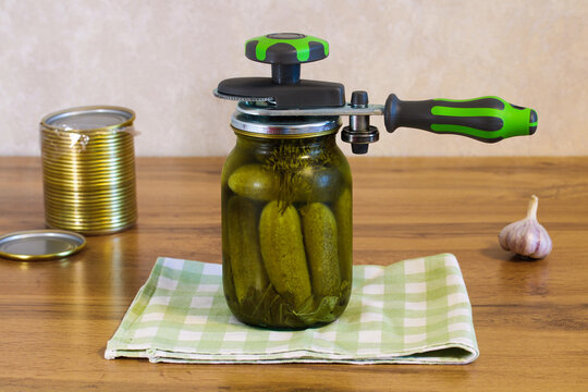 Glass jar filled with homemade pickled cucumbers and herbs, sealed with a manual canning machine on a wooden kitchen table with garlic