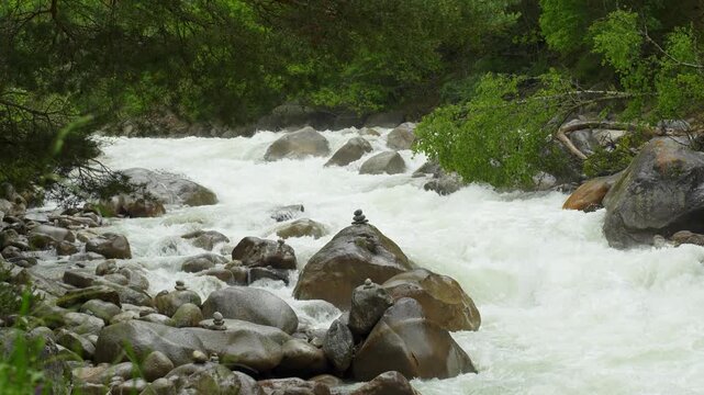 A dynamic section of a mountain river: foaming water rushes between massive, rounded boulders, curving around them and forming eddies. In the foreground are neatly stacked pyramids of stones. Along th