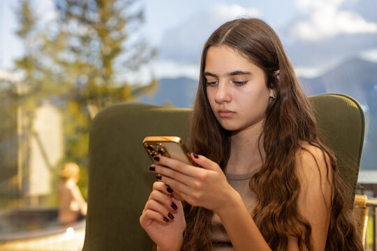 Teenage Girl Using Smartphone While Seated by a Window. Teenage girl with long brown hair sits in a green chair using a smartphone, with trees and blurred mountains visible through the window.