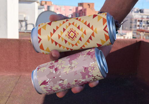 Mockup of hand holding two blank cans on a rooftop with buildings in the background. It is daytime and the sky is clear.