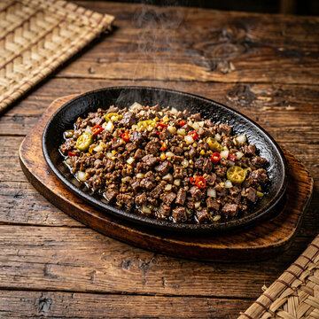 Sizzling chopped meat with onions, red chilies and citrus served steaming on a hot cast iron platter over a rustic wooden table.