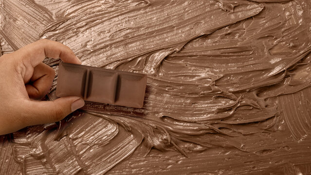 Hand holding a milk chocolate bar piece over a textured background of spread chocolate cream. World Chocolate Day celebration