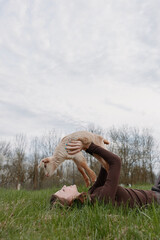Naklejka premium Young woman lies on grass in a brown sweater lifting a baby goat above her with a joyful expression in a natural outdoor setting under cloudy sky