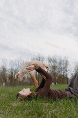 Naklejka premium Young woman lies on green grass holding a baby goat above her with joy in a natural outdoor setting under cloudy sky