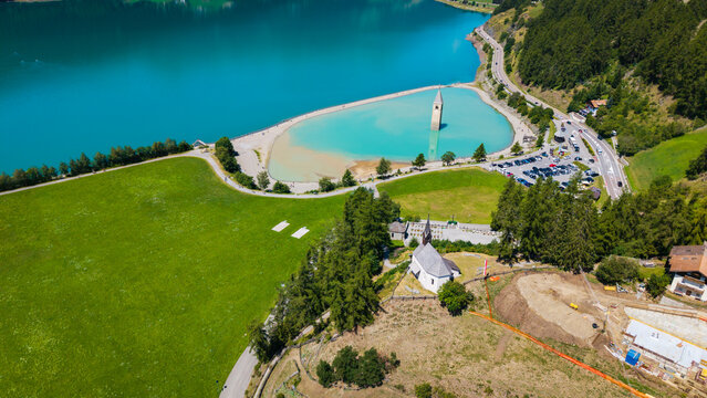 aerial drone view of Lake Resia submerged bell tower and surrounding green meadows along the shoreline in Curon Venosta, South Tyrol, Italy.