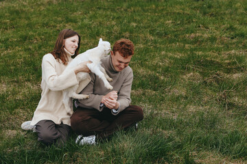 Naklejka premium Young couple enjoys playful moment with a white baby goat while sitting on green grass in casual sweaters during a relaxed lifestyle outdoor scene.
