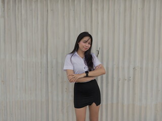 Young Asian woman in a short-sleeved white button-up shirt and black mini skirt stands in front of a light corrugated metal partition © iFocus