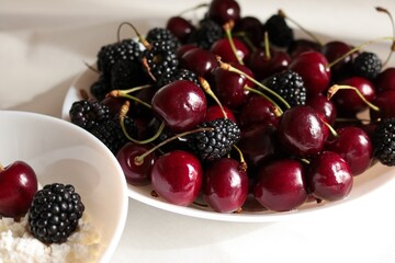 Close-up of a white plate filled with fresh ripe sweet cherries and blackberries with cottage cheese bowl in morning light. © Svetlana