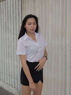 Young Asian woman in a short-sleeved white button-up shirt and black mini skirt stands in front of a light corrugated metal partition