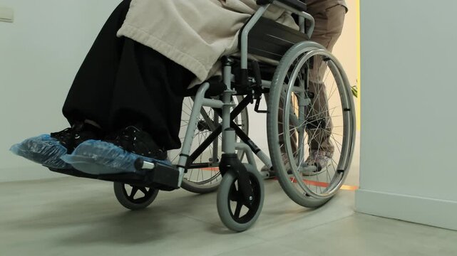 Nurse helps paralyzed patient move safely through sterile clinical hallway. Doctor pushes wheelchair past hospital doors. Slow motion