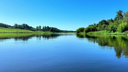 Fototapeta premium Mirror-like river surface reflecting lush green banks under a vibrant blue sky