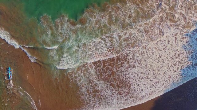 Aerial view of a small boat on the sandy shore of Baron Beach with crashing waves in Yogyakarta, Special Region of Yogyakarta, Indonesia.