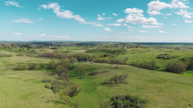 Aerial view over green prairie grassland and rolling hills near Mandan, North Dakota, with a small pond, brushy draws and scattered trees under a bright spring sky.