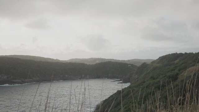 Birds flying over a green peninsula and a calm bay on an overcast day in Buzios, Brazil