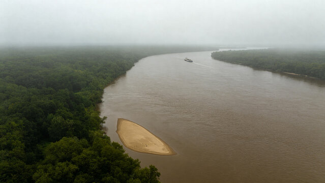 View from high bluff of wide brown river bending through low green forest, sandbar, tugboat and barge downstream, Mississippi River landscape