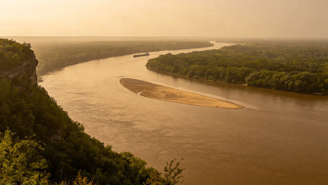 View from high bluff of wide brown river bending through low green forest, sandbar, tugboat and barge downstream, Mississippi River landscape