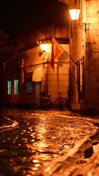 Narrow old stone street of Mediterranean coastal town during rainy night with glowing street lamps and parked vintage bicycle.