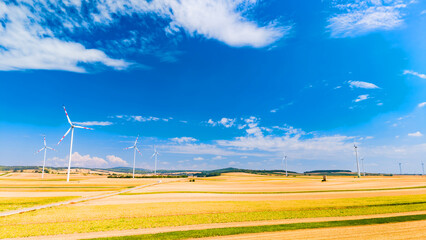 Wind turbines stand in open fields under a bright blue sky. Wind turbines rotate in a flat landscape with fields during sunny weather. Clouds drift above the horizon. © Vadim