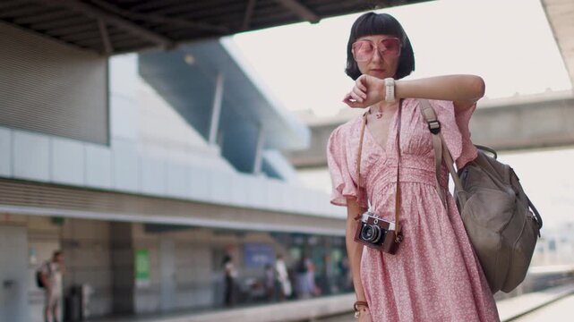A female traveler with a camera and a backpack stands on a railway station platform. She is checking her watch and waiting for the train. Train travel. A young woman with a camera.