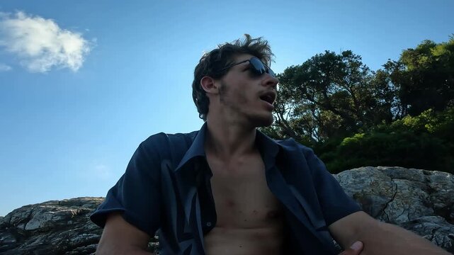 Confident male vlogger in unbuttoned shirt relaxing on rocky beach and recording video message for social media vlog.