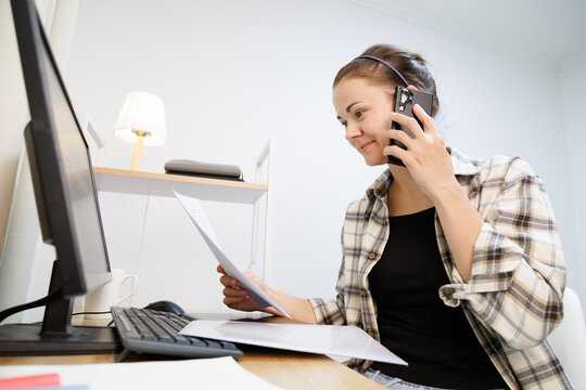 Talking on the phone while checking documents at a computer desk, handling paperwork, business communication and daily office tasks from home.