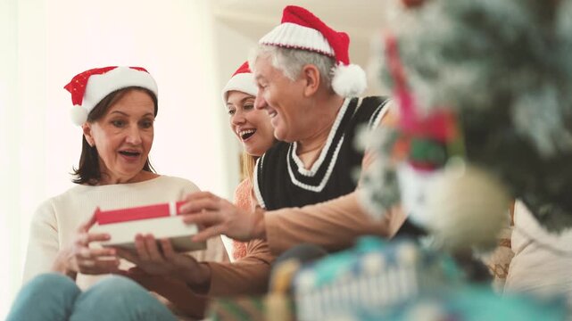 Adult daughter hugging senior parents while father gives mother Christmas present, family celebration, gift exchange, holiday surprise, multigenerational Christmas at home.