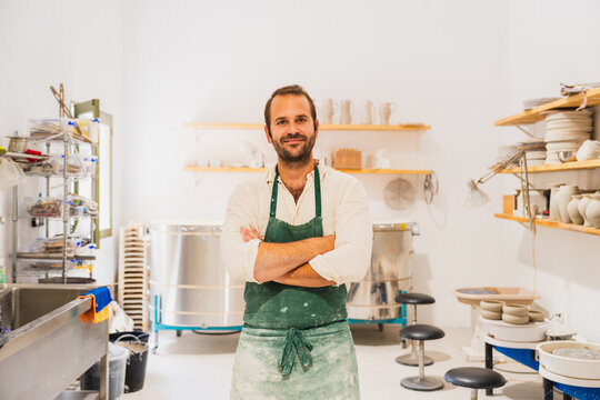 Smiling male ceramist with apron standing in his pottery workshop
