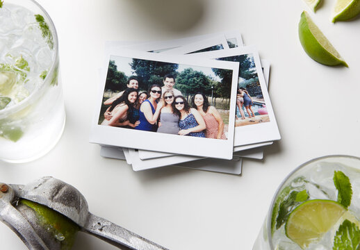 On a table, there are several blank polaroid frames stacked together. Nearby, there are drinks with ice and lime slices. PSD template mockup