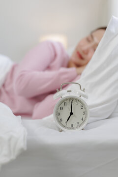 Alarm clock showing seven o'clock beside sleeping woman in bed, representing morning wake-up time, sleep routine, peaceful rest, bedroom comfort, relaxation and daily schedule.