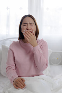 Sleepy woman yawning after waking up in bed, covering her mouth with hand in a bright bedroom, representing morning fatigue, tiredness, drowsiness, sleepiness, bedtime routine and lack of sleep.
