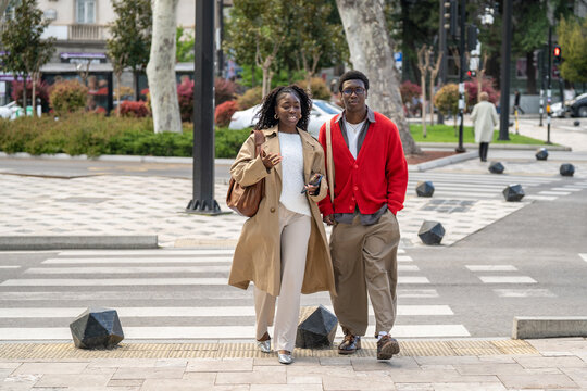 Fashionable young african american couple man and woman close friends meeting for daily walk in city, positive black male and female date walking on crosswalk chatting and sharing latest news.
