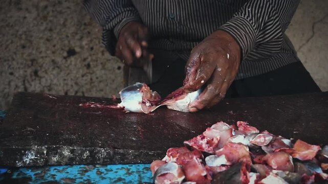 Close-up of a person's hands expertly chopping fresh fish into pieces on a dark cutting board, preparing seafood for sale in a bustling market.
