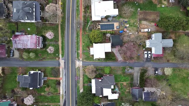 Vibrant red brick pedestrian sidewalk parallels Washington Street across paved crossroads. Blooming spring dogwood trees shade historic tin roof homes in Harpers Ferry, West Virginia