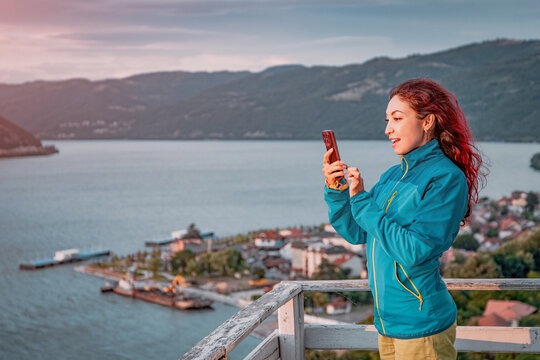 Young woman framed against a sweeping river panorama photographs the scenic view below from a hilltop overlook. Travel concept.