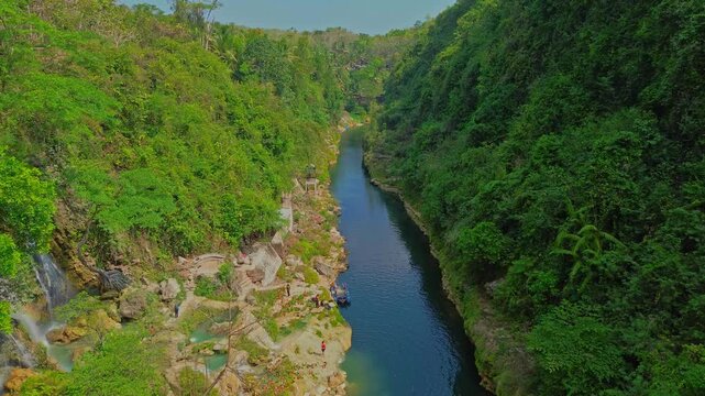 Aerial view of Sri Gethuk Waterfall flowing into a calm river surrounded by lush green tropical forest and rocky terrain in Yogyakarta, Special Region of Yog, Indonesia.
