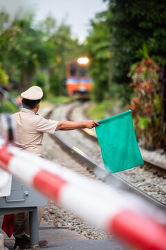 Selective focus to Thai railway officer holding a green signal flag at a level crossing safety barrier as a passenger train approaches along the tracks in a lush green environment.