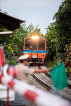 Selective focus to passenger train with blurry Thai railway officer holding a green signal flag at a level crossing safety barrier as a passenger train approaches along the tracks in a lush green .