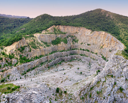 Aerial view of an abandoned open-pit stone quarry with terraced rock walls surrounded by green forested hills.