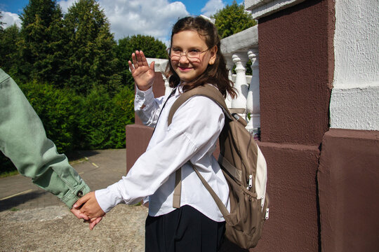 A schoolgirl stands outside her school waving with a smile while holding hands with her mother. The setting is bright and sunny