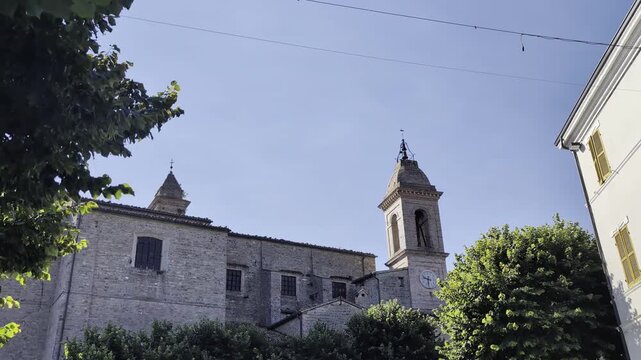 La suggestiva veduta di una chiesa storica italiana in pietra con campanile, circondata da alberi verdi sotto un cielo azzurro e limpido