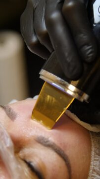 Professional beautician applying cleansing mask on female client's face. Close-up of female face during youth restoration and facial rejuvenation procedure  - Vertical Video