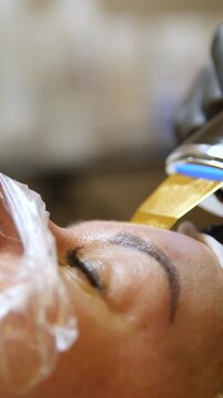 Professional beautician applying cleansing mask on female client's face. Close-up of female face during youth restoration and facial rejuvenation procedure  - Vertical Video