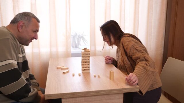 Two players take turns removing wooden blocks from a tower while concentrating on a tabletop game at home.