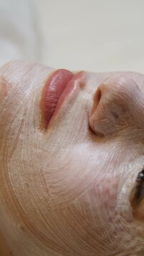 Professional beautician applying cleansing mask on female client's face. Close-up of female face during youth restoration and facial rejuvenation procedure  - Vertical Video